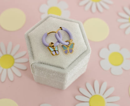 A pair of kids small hypoallergenic butterfly hoop earrings with lavender purple hoops and gold-plated butterfly charms featuring mint green and lavender enamel, displayed on a white box against a soft pink background with flowers. Earrings are from the Pierced Peanut Earring co in Nz