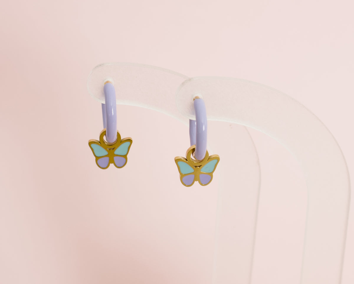 A pair of kids small hypoallergenic butterfly hoop earrings with lavender purple hoops and gold-plated butterfly charms featuring mint green and lavender enamel, displayed on a clear acrylic stand against a soft pink background. Earrings are from the Pierced Peanut Earring co in Nz