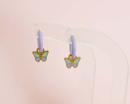A pair of kids small hypoallergenic butterfly hoop earrings with lavender purple hoops and gold-plated butterfly charms featuring mint green and lavender enamel, displayed on a clear acrylic stand against a soft pink background. Earrings are from the Pierced Peanut Earring co in Nz