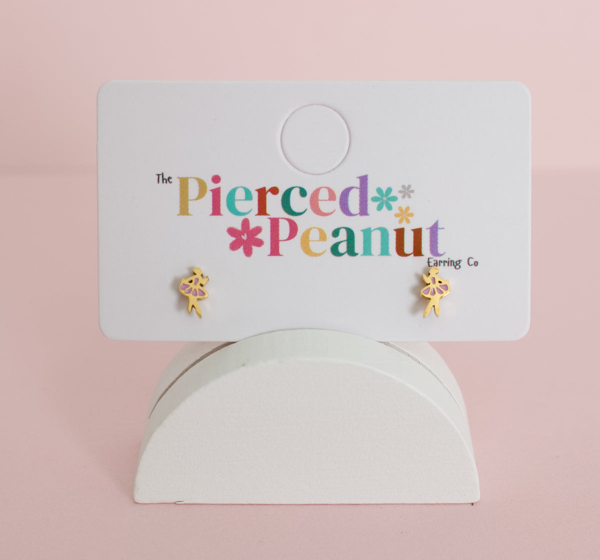 A pair of kids small hypoallergenic gold-tone ballerina screwback stud earrings displayed on a white branded card that reads 'The Pierced Peanut Earring Co,' set against a soft pink background.
