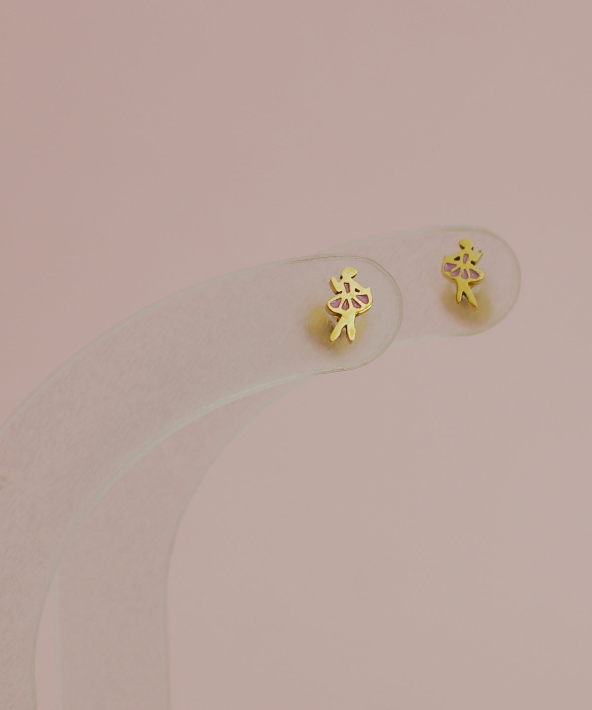 A pair of kids small hypoallergenic gold screwback stud earrings shaped like ballerinas with pink accents, displayed on a clear curved stand against a soft dusty pink background. Earrings are from the brand the Pierced Peanut earring co in NZ.
