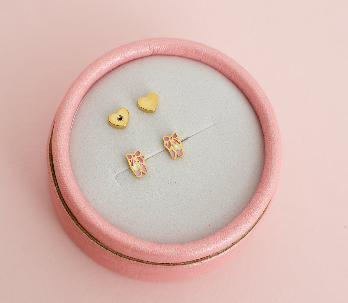 A pair of kids hypoallergenic gold ballet shoe stud earrings displayed inside a round pink jewellery box. The earrings are from the Pierced Peanut earring co in New Zealand.