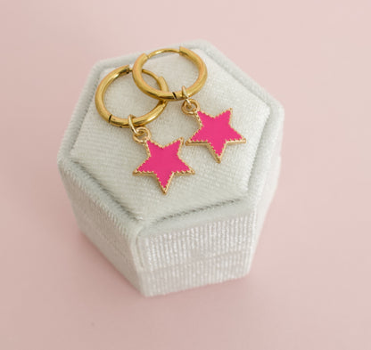 A pair of kids hypoallergenic gold huggies with pink star charms. Displayed on a white box with a soft pink background by the Pierced Peanut earring co in Nz