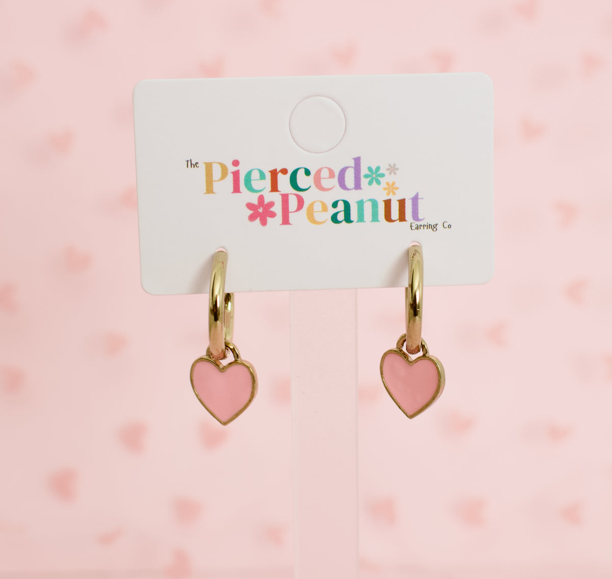 Two gold-tone hypoallergenic hoop earrings with pink heart charms, displayed on a white branded jewellery card reading The Pierced Peanut Earring Co, based in New Zealand.