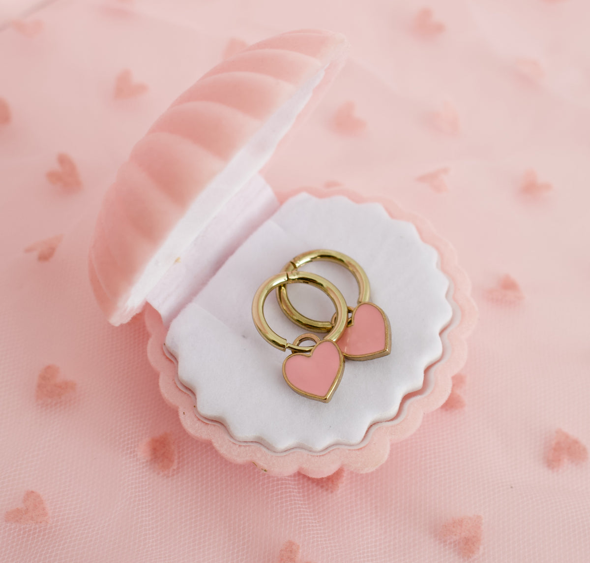 Two gold-tone hypoallergenic hoop earrings with pink heart charms, displayed inside a pink and white scalloped jewelry box on a pink heart-patterned fabric background. The earrings are from the brand the Pierced Peanut in NZ.