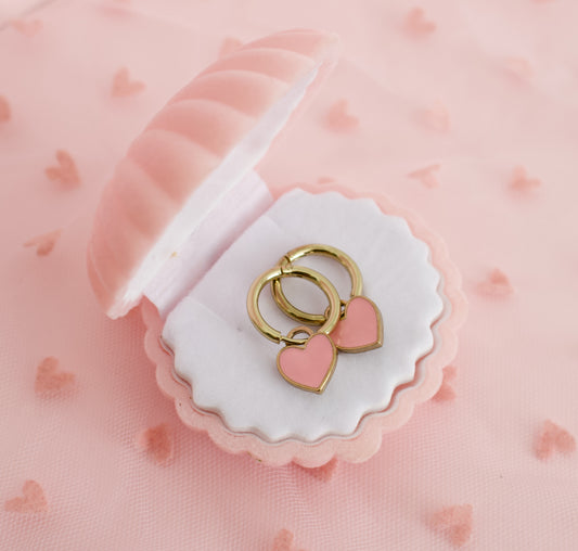 Two gold-tone hypoallergenic hoop earrings with pink heart charms, displayed inside a pink and white scalloped jewelry box on a pink heart-patterned fabric background. The earrings are from the brand the Pierced Peanut in NZ.