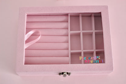A pink felt jewellery organizer box with a hinged lid, featuring a large roll-top compartment, a side grid of compartments, and a small silver clasp. A pink ribbon is tucked into the roll-top compartment and the box is from The Pierced Peanut Earring Company in Nz
