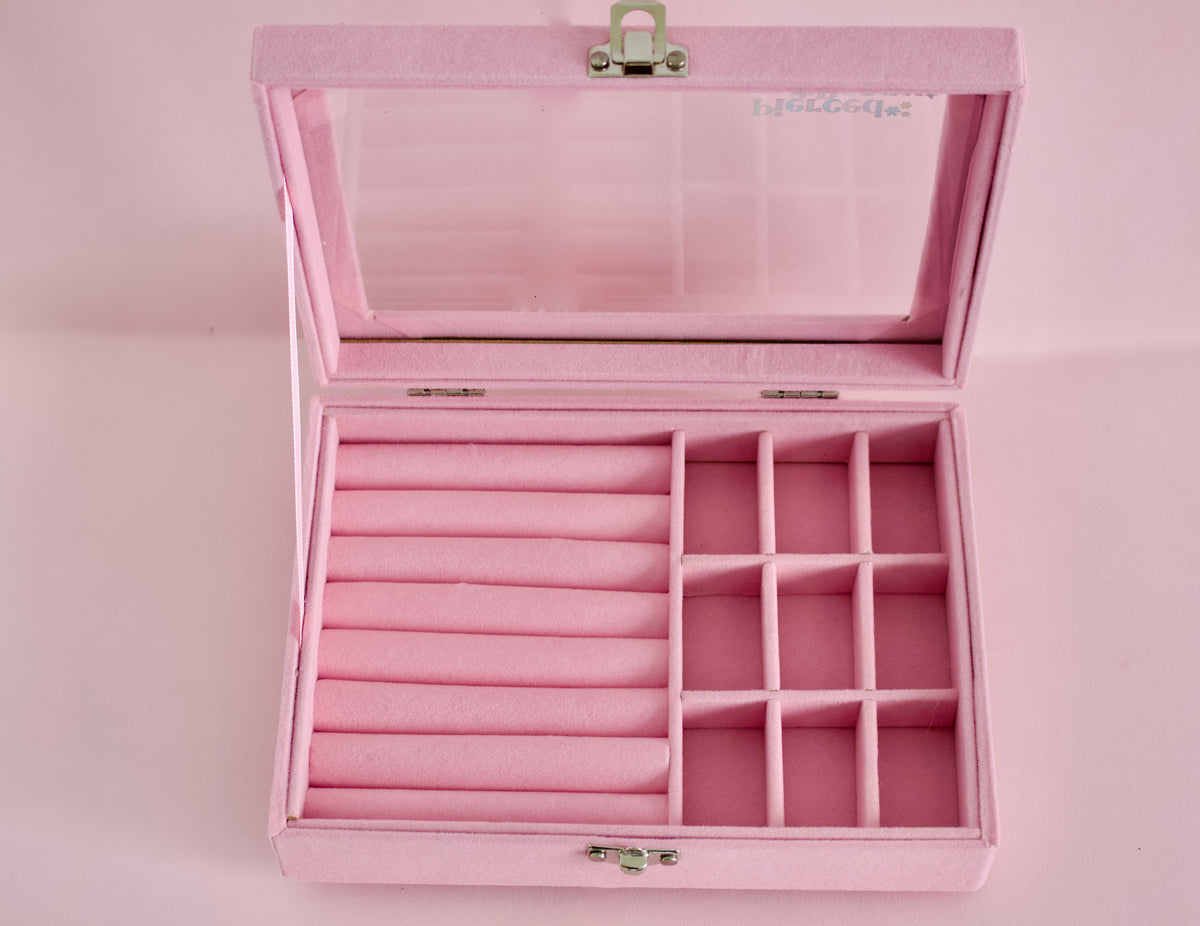 An open pink velvet jewelry box with a hinged lid, featuring a large roll-top section on the left and multiple compartment sections on the right, set against a solid pink background from The Pierced Peanut in NZ.