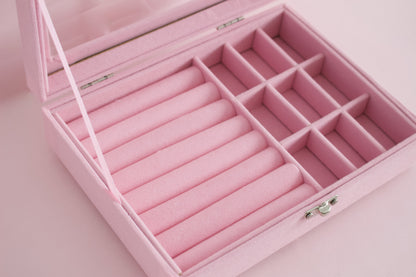 A pink velvet jewelry organizer box with a hinged lid, featuring cylindrical ring slots and a grid of small compartments, shown open on a matching pink background from the Pierced Peanut Earring Co in New Zealand.