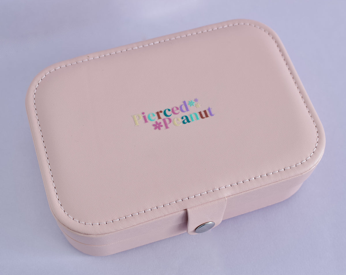 A light pink faux-leather jewellery organizer box with a snap button closure, featuring the text 'Pierced* Peanut' in a colourful gradient font on the lid from NZ.