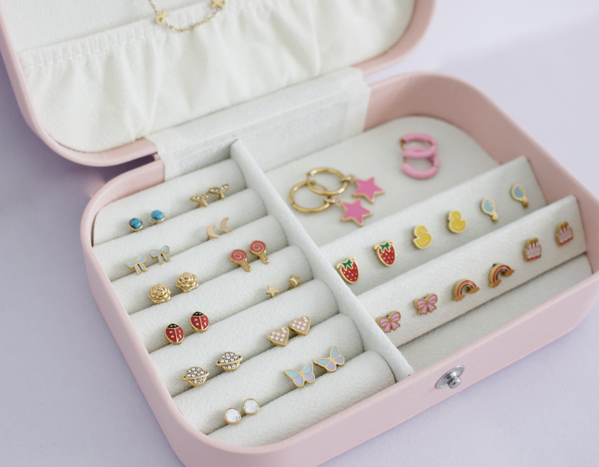 A pink jewelry box with a cream velvet interior, open to display multiple pairs of colorful stud earrings arranged in compartments. The earrings feature playful shapes including strawberries, butterflies, rainbows, stars, and hearts in gold, pink, red, blue, and white. From the Pierced Peanut earring co in Nz.