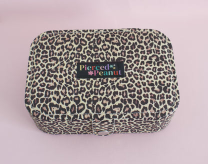 A leopard print fabric jewellery box with a black label reading 'Pierced by Peanut' in colourful text, set against a solid light pink background.