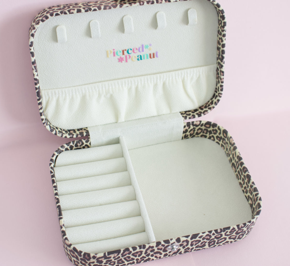 An open leopard-print jewellery organiser case with cream-coloured interior compartments, including hook slots on the lid and a ring roll, branded 'Pierced * Peanut' from NZ in pastel text on the inside lid, set against a solid pink background.