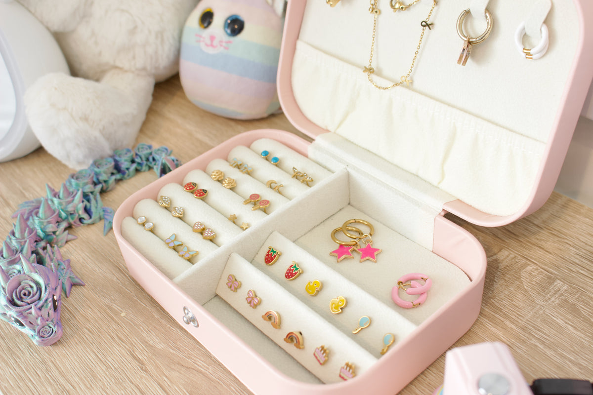 A pink hard-shell jewellery organizer box shown open on a wooden surface, filled with rings, earrings, and small jewellery pieces. A plush toy and pastel decorative items are visible in the background. From the Pierced Peanut Earring Co in New Zealand.