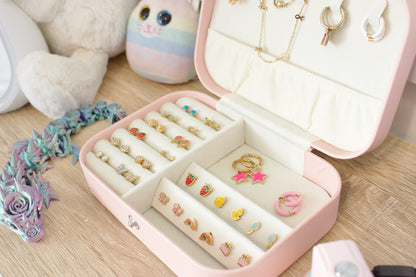 A pink hard-shell jewellery organizer box shown open on a wooden surface, filled with rings, earrings, and small jewellery pieces. A plush toy and pastel decorative items are visible in the background. From the Pierced Peanut Earring Co in New Zealand.