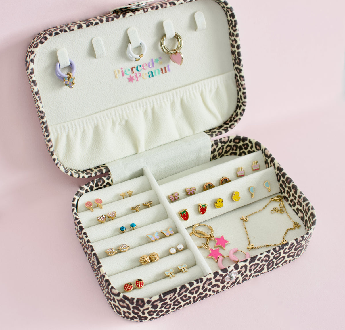 An open leopard-print jewellery organiser case filled with earrings, rings, and a necklace, photographed on a soft pink background. The inside lid has ring slots and a small multicolor 'Pierced by Peanut' text detail in New Zealand.