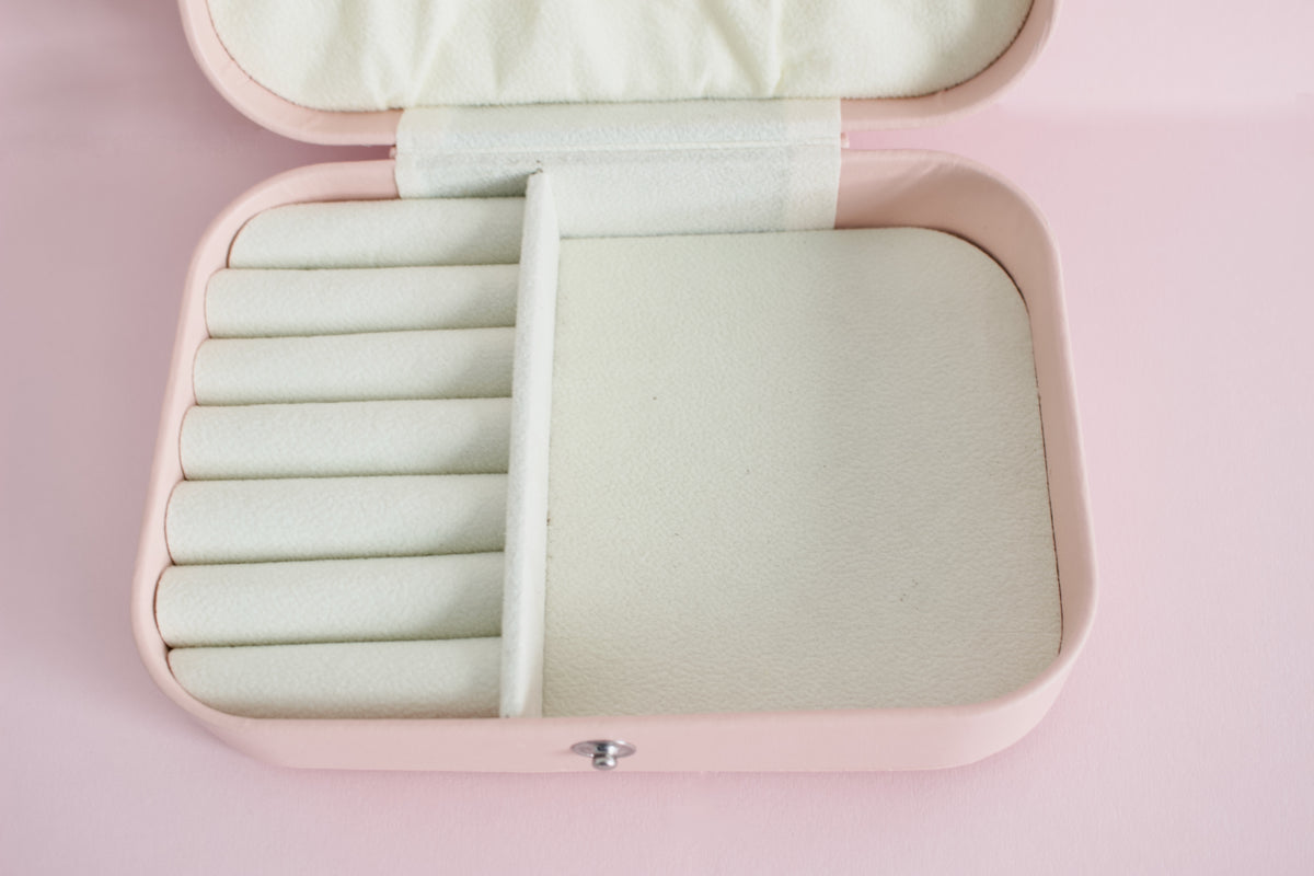 An open pink jewellery box with a cream-coloured interior, featuring a ring roll on the left and a larger flat compartment on the right, set against a solid pink background from the Pierced Peanut in NZ.