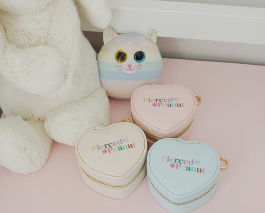 Three pastel heart-shaped zipper jewellery travel pouches with gold zippers and the text 'Pierced Peanut' in rainbow lettering, displayed on a pink surface next to a white plush toy and a cat-face plush.