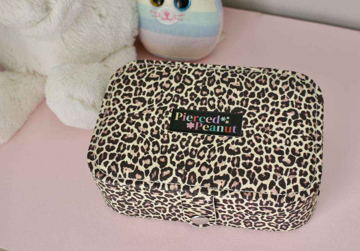 A leopard print jewellery box with a black brand label reading 'Pierced * Peanut' sits on a pink surface next to a white plush toy and a pastel plush character from New Zealand.