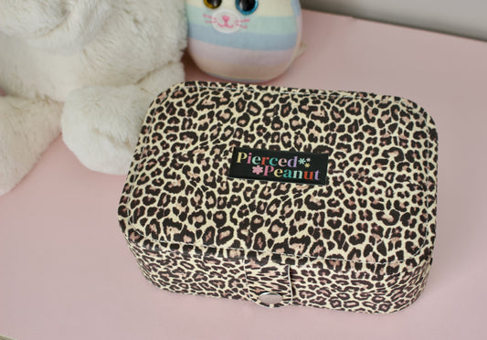 A leopard print jewellery box with a black brand label reading 'Pierced * Peanut' sits on a pink surface next to a white plush toy and a pastel plush character from New Zealand.