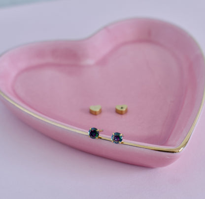 A pair of small gold stud earrings with dark blue iridescent gemstones resting on the edge of a pink heart-shaped ceramic dish with a gold rim, with a second pair of gold heart studs inside the dish from the Pierced Peanut Earring Co in Nz