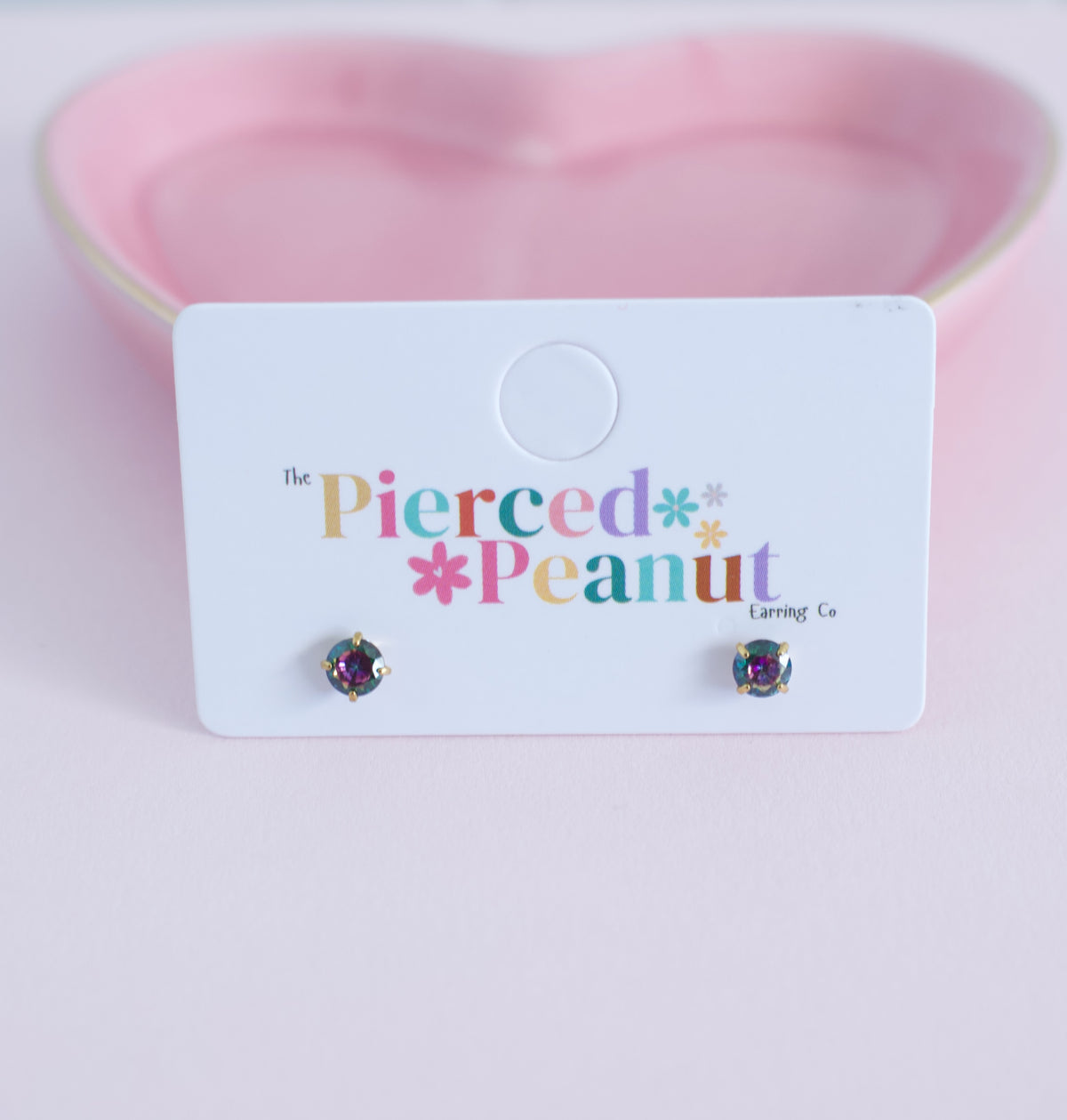 A pair of small round stud earrings with iridescent purple and green stones, displayed on a white card that reads 'The Pierced Peanut Earring Co' against a soft pink heart-shaped background.