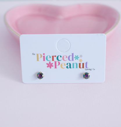 A pair of small round stud earrings with iridescent purple and green stones, displayed on a white card that reads 'The Pierced Peanut Earring Co' against a soft pink heart-shaped background.