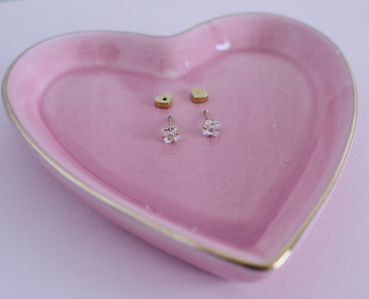 A pair of small gold stud earrings displayed on a pink heart-shaped ceramic tray with a gold rim, set against a soft pink background from the pierced peanut earring co in nz.
