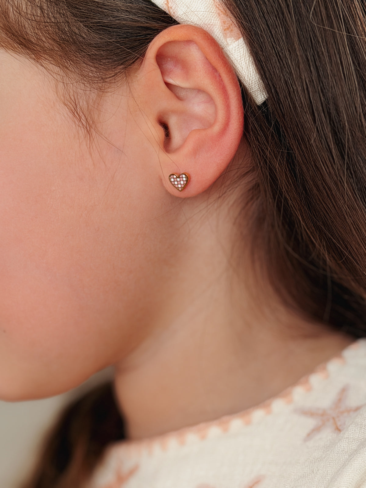 A close-up side profile of a girl wearing a small heart-shaped stud earring with a textured, glittery rose gold surface from the pierced peanut earring co.