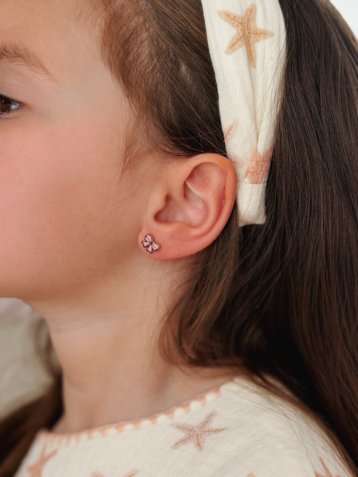 A close-up side profile of a young girl wearing a small hypoallergenic pink bow stud earring. The earrings are screwback studs from the Pierced Peanut Earring Co in Nz.