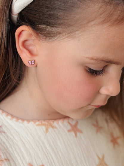 A close-up of a young girl wearing a small hypoallergenic pink bow stud earring looking down. The earrings are screwback studs from the Pierced Peanut Earring Co in Nz.