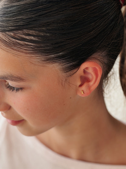 A close-up side profile of a young girl with dark hair tied back, wearing a tiny gold ribbon stud earring on her earlobe. The earring is a hypoallergenic screwback stud from The Pierced Peanut Earring Co in Nz in collaboration with the Child Cancer Foundation Nz