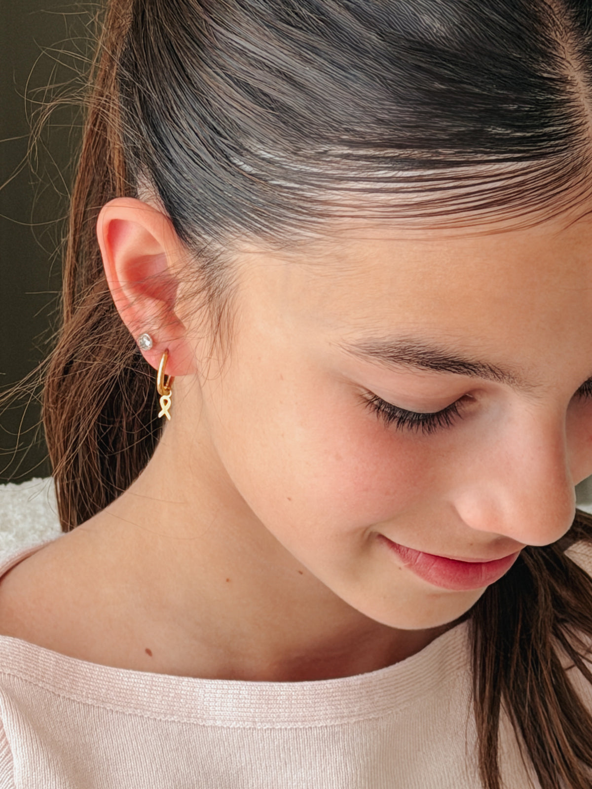 A close-up side profile of a young girl with dark hair tied back, wearing a tiny gold ribbon huggie earring on her earlobe. The earring is a hypoallergenic gold hoop earring from The Pierced Peanut Earring Co in Nz in collaboration with the Child Cancer Foundation Nz