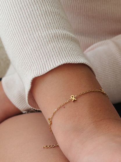 A delicate gold chain bracelet with a small gold ribbon charm worn on a person's wrist, paired with a ribbed cream-colored top. The bracelet is hypoallergenic and waterproof from the Pierced Peanut Earring Co in Nz in collaboration with the Child Cancer Foundation Nz.