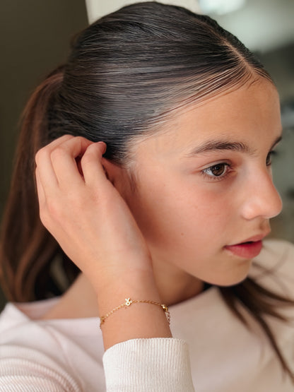 A close-up lifestyle shot of a young girl with dark hair in a ponytail, wearing a delicate gold chain bracelet with a small gold ribbon charm on her wrist. The bracelet is hypoallergenic and waterproof from the Pierced Peanut Earring Co in Nz in collaboration with the Child Cancer Foundation Nz