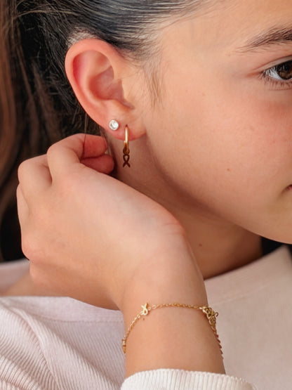 A close-up lifestyle shot of a model wearing a delicate gold huggie hoop earring with a dangling ribbon charm, along with a matching gold ribbon bracelet. The earring and bracelet are hypoallergenic from the Pierced Peanut Earring Co in collaboration with the Child Cancer Foundation in Nz.