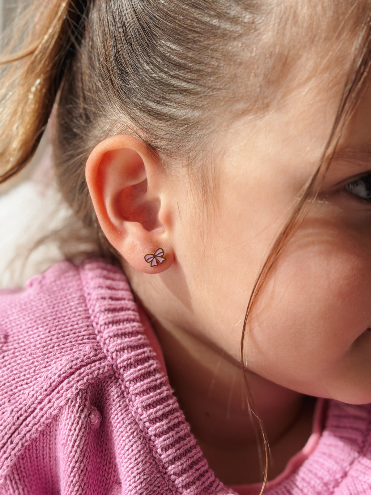 A close-up side profile of a young girl wearing a small hypoallergenic pink bow stud earring. The earrings are screwback studs from the Pierced Peanut Earring Co in Nz.