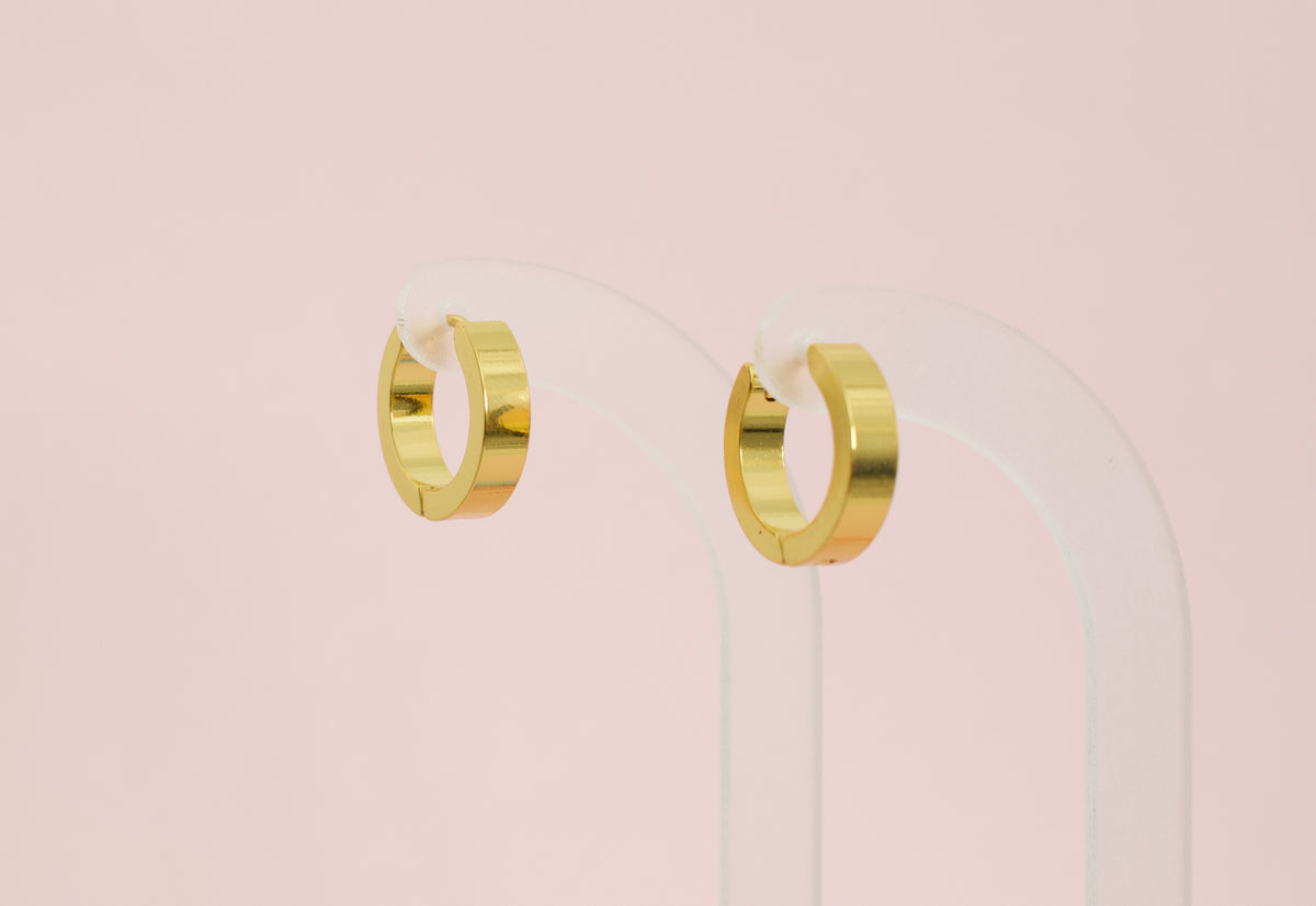 A pair of kids hypoallergenic polished gold huggie hoop earrings displayed on a clear acrylic stand against a soft pink background. The earrings are from the brand The Pierced Peanut Earring Co in New Zealand, NZ.