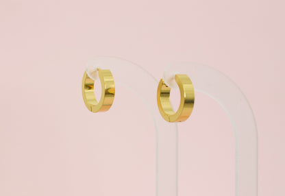 A pair of kids hypoallergenic polished gold huggie hoop earrings displayed on a clear acrylic stand against a soft pink background. The earrings are from the brand The Pierced Peanut Earring Co in New Zealand, NZ.