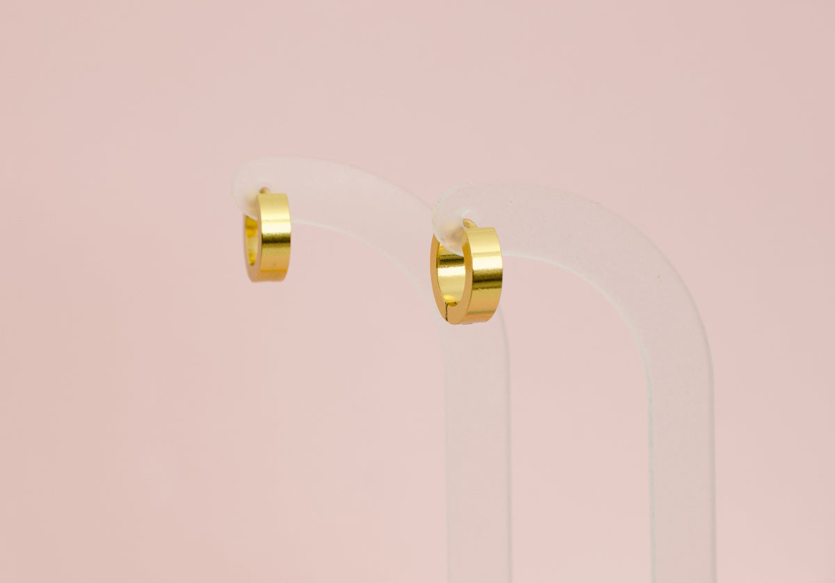 A pair of kids hypoallergenic polished gold huggie hoop earrings displayed on a clear acrylic stand against a soft pink background. The earrings are from the brand The Pierced Peanut Earring Co in New Zealand, NZ.