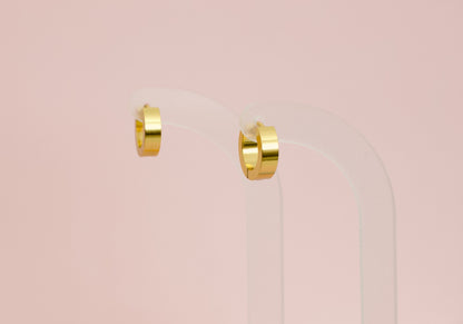 A pair of kids hypoallergenic polished gold huggie hoop earrings displayed on a clear acrylic stand against a soft pink background. The earrings are from the brand The Pierced Peanut Earring Co in New Zealand, NZ.