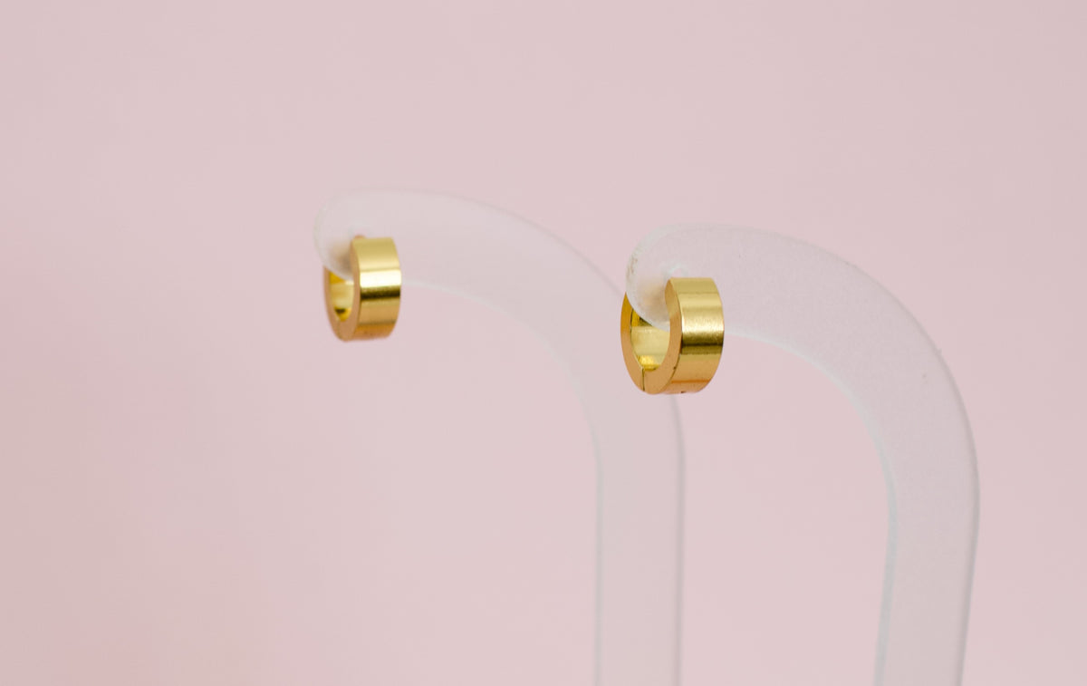 A pair of kids hypoallergenic polished gold huggie hoop earrings displayed on a clear acrylic stand against a soft pink background. The earrings are from the brand The Pierced Peanut Earring Co in New Zealand, NZ.