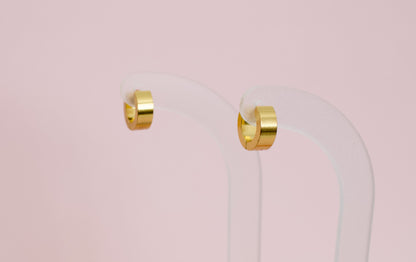 A pair of kids hypoallergenic polished gold huggie hoop earrings displayed on a clear acrylic stand against a soft pink background. The earrings are from the brand The Pierced Peanut Earring Co in New Zealand, NZ.